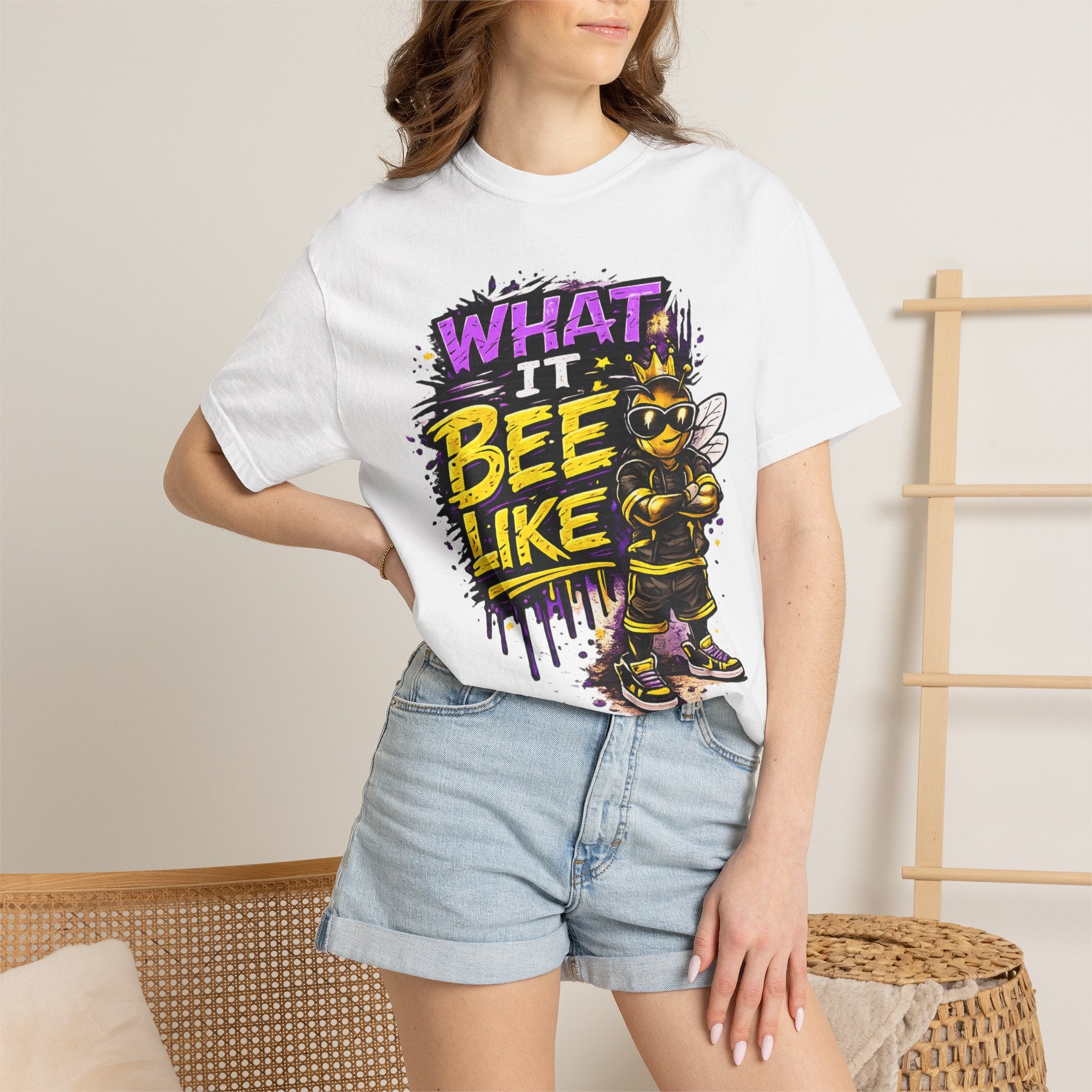 A woman wearing a white graphic t-shirt with a bee-themed print that reads 'WHAT IT BEE LIKE' in purple and yellow, paired with light blue denim shorts, in a brightly lit indoor setting.